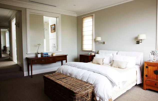 Modern bedroom with white bedding, wicker trunk, wooden furniture, large window, and wall sconces.