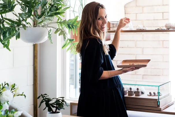 A woman in a black dress holds a tray of jewelry, standing in a bright room with plants and a display case.