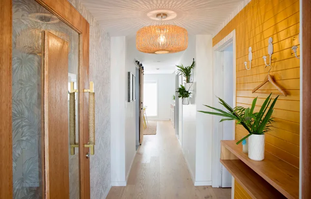 Hallway with wooden accents, tropical wallpaper, and plants; bright lighting from a woven lampshade casts shadows.