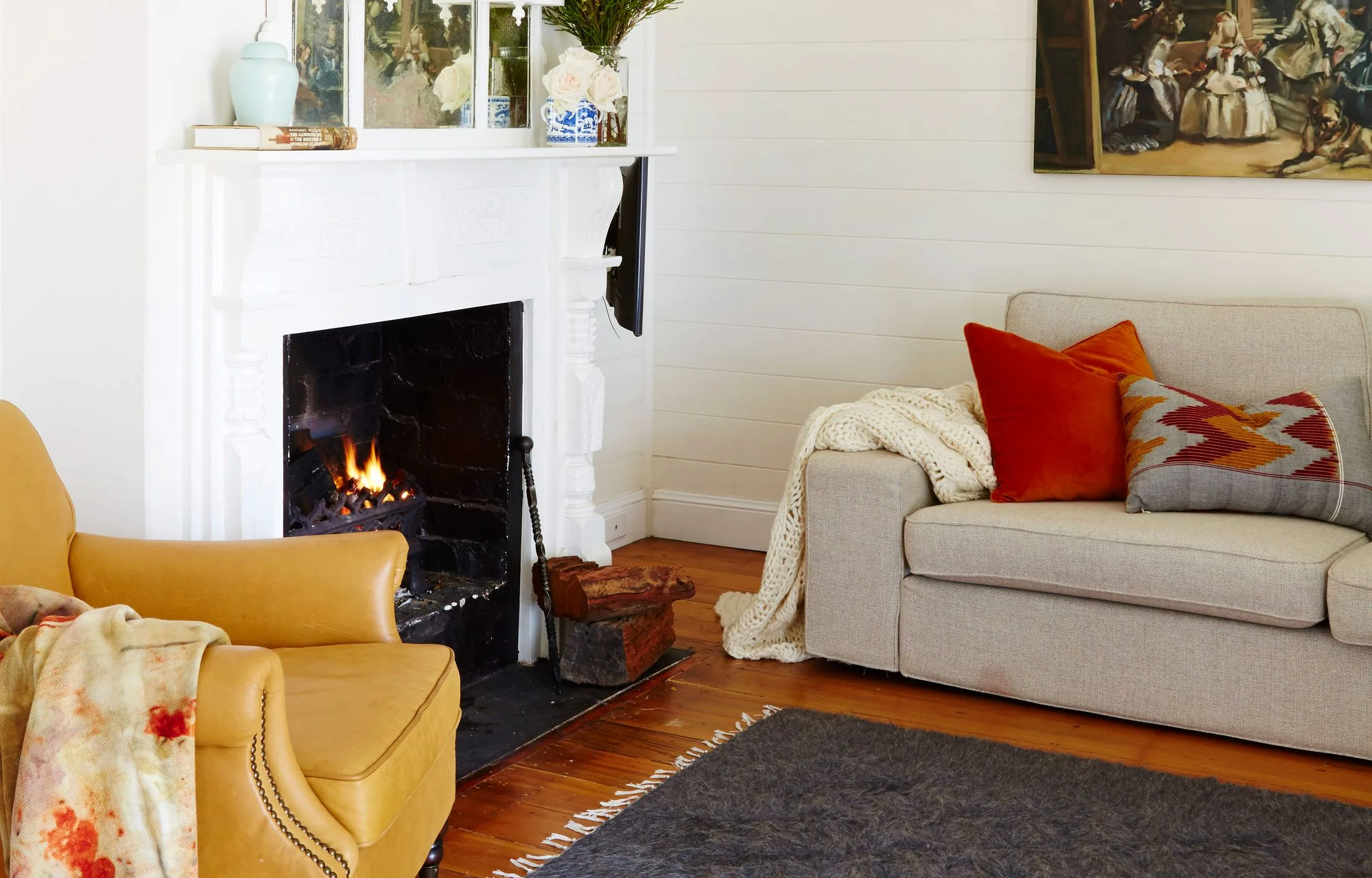Cozy living room with a lit fireplace, beige sofa, orange pillows, and an armchair, featuring warm decor elements.