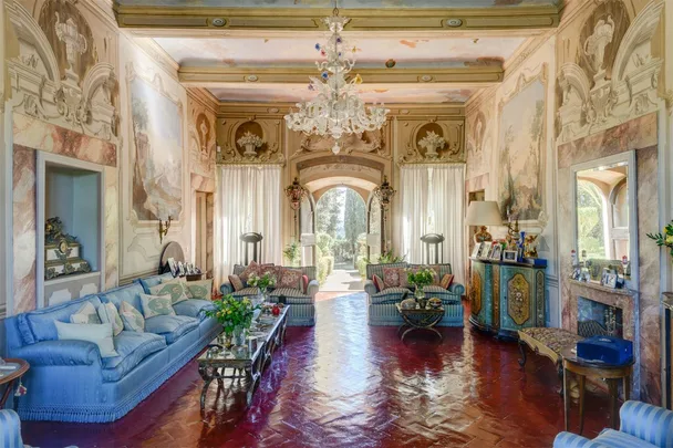 Elegant living room with ornate ceilings, large blue couch, patterned chairs, and a central chandelier.
