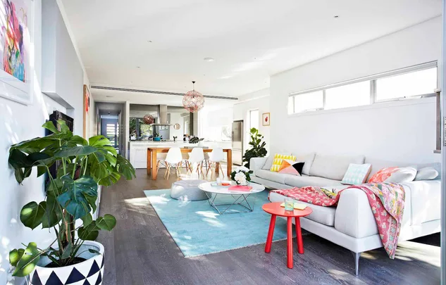 Modern living room with white sofa, colorful cushions, red side table, indoor plants, and dining area in the background.