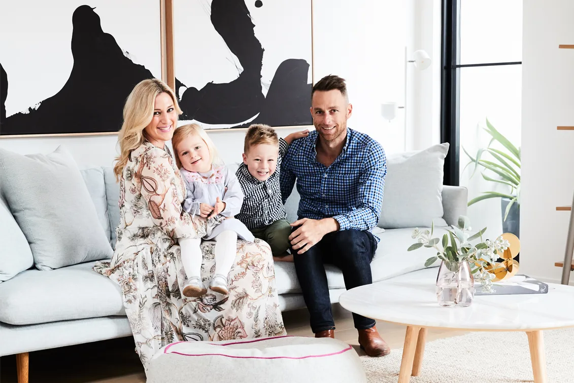 A family of four smiling and sitting on a sofa in a modern, well-lit living room with abstract art on the wall.