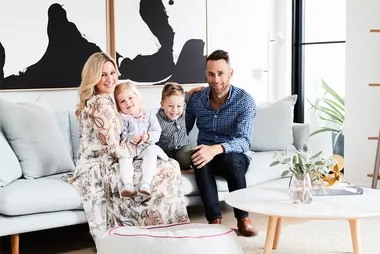 A family of four smiling and sitting on a sofa in a modern, well-lit living room with abstract art on the wall.