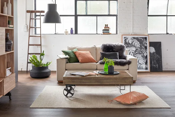 Cozy living room with beige sofa, colorful cushions, wooden coffee table, plants, artwork, and loft-style windows.
