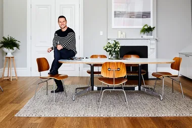 Man in striped sweater sitting on table in modern dining room with wooden chairs, plants, and a fireplace in the background.