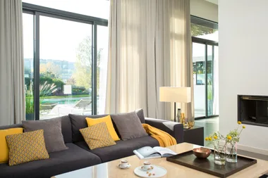 Living room with large windows, gray sofa, yellow cushions, coffee table with vase, and view of garden.