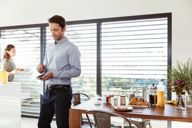 Man in blue shirt using smartphone, standing near a table with breakfast items, woman in background holding a tray.