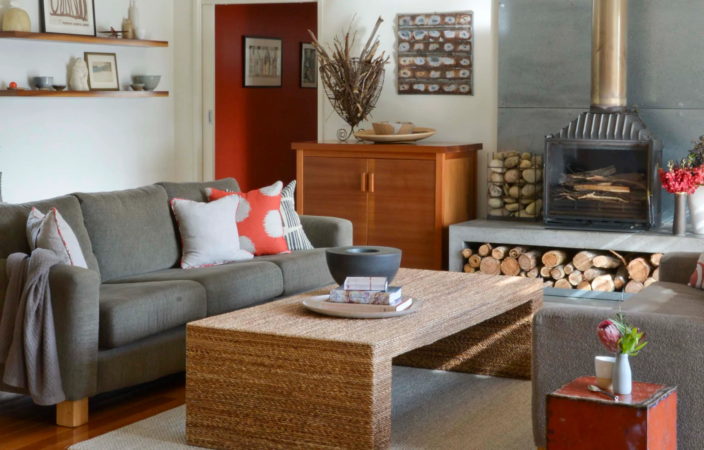 Cozy living room with a grey sofa, colorful pillows, woven coffee table, fireplace, and decorative elements.