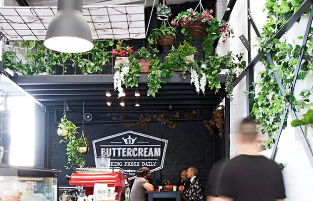 Indoor cafe scene with hanging plants, people sitting, and a sign reading "Buttercream Baking Fresh Daily."