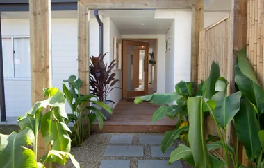 Outdoor entryway with wooden door, surrounded by lush green plants, leading into a modern corridor.