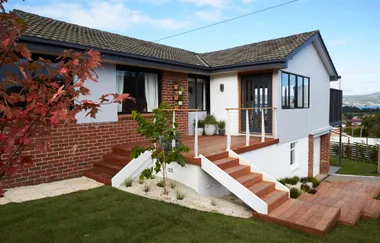 A brick house with a wooden deck, stairs, and garden, set against a blue sky.