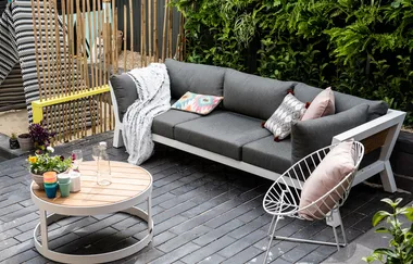 Outdoor patio with a gray sofa, patterned pillows, round table, colorful cups, and greenery in the background.