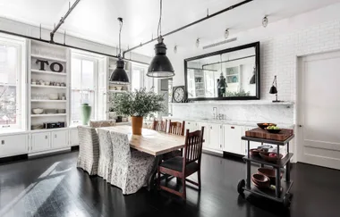 Spacious kitchen with a wooden table, chairs, large windows, hanging lamps, and white cabinets with marble countertops.