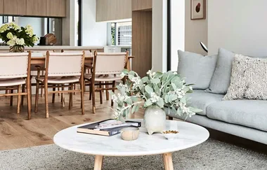 Living room with a white round table, vase of flowers, gray sofa, and wooden dining chairs in the background.