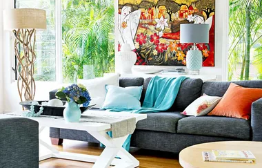 Stylish living room with a gray sofa, colorful cushions, a floral painting, and large windows showing green plants outside.