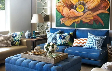 Bright living room with blue sofa, colorful cushions, a large flower painting on the wall, and a tufted ottoman.