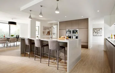 Modern kitchen with island, bar stools, pendant lights, and built-in appliances. Dining area visible in the background.