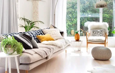 Cozy living room with a beige sofa, assorted cushions, plants, a chair, and large window bringing in natural light.