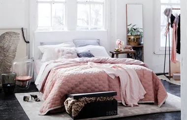 Cozy bedroom with pink bedding, fluffy pillows, stylish decor, and a trunk labeled 1862 at the foot of the bed.