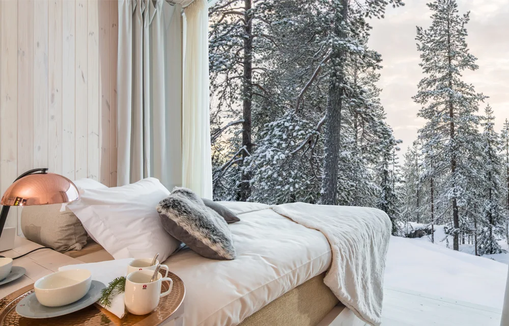 Cozy bedroom with a view of snow-covered pine trees, featuring a bed, pillows, and a copper lamp.