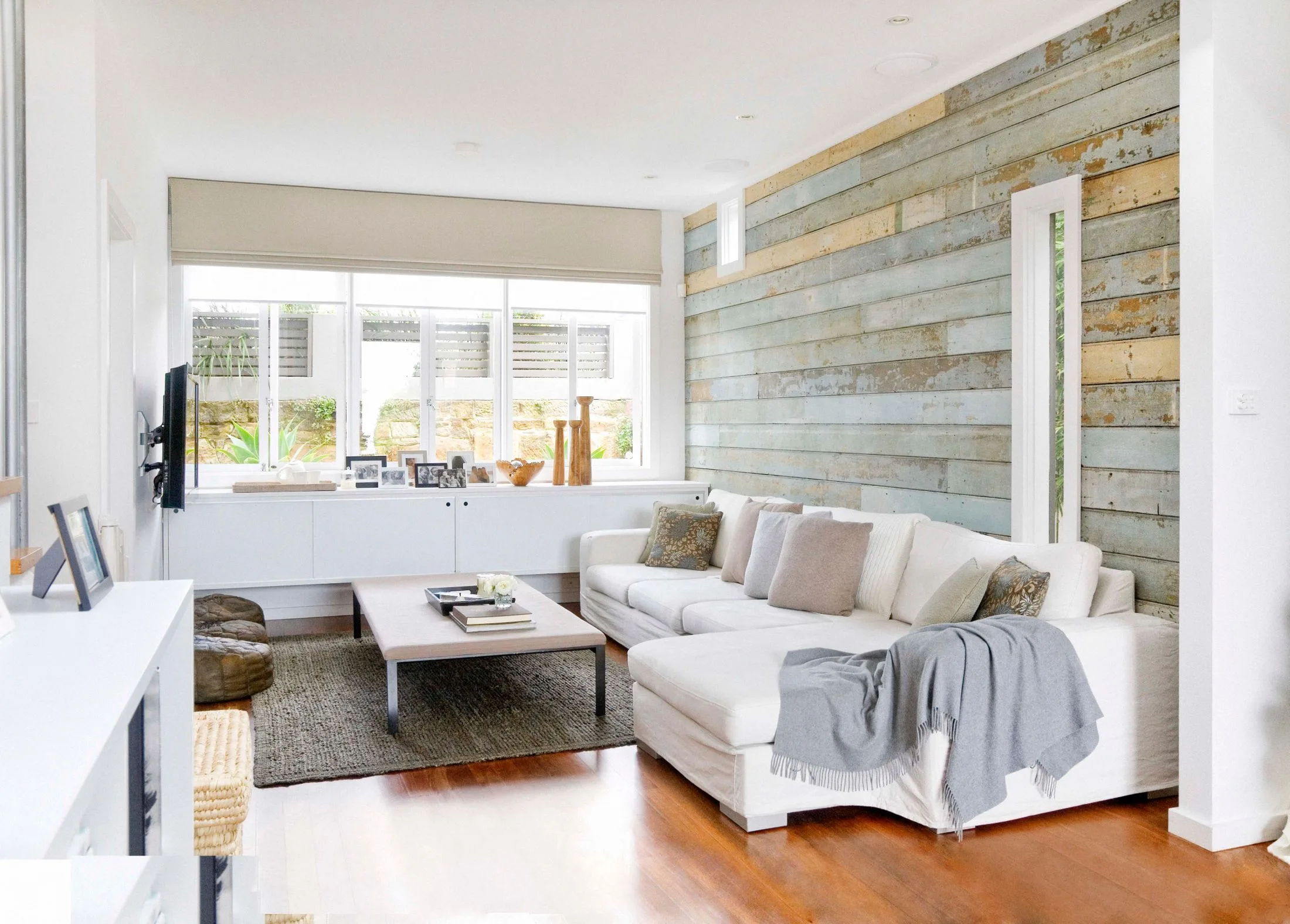 Modern living room with white sofas, rustic wood accent wall, large windows, and a coffee table with decor items.