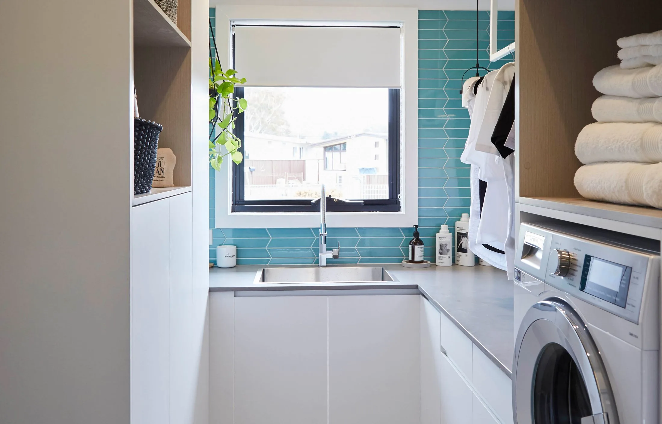 Laundry room with a window, teal tiles, sink, plants, washer, shelves with towels and detergent bottles.