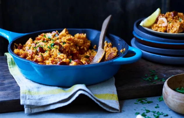 A blue skillet filled with colorful paella, featuring rice, chorizo, and herbs, on a wooden board with a wooden spoon.