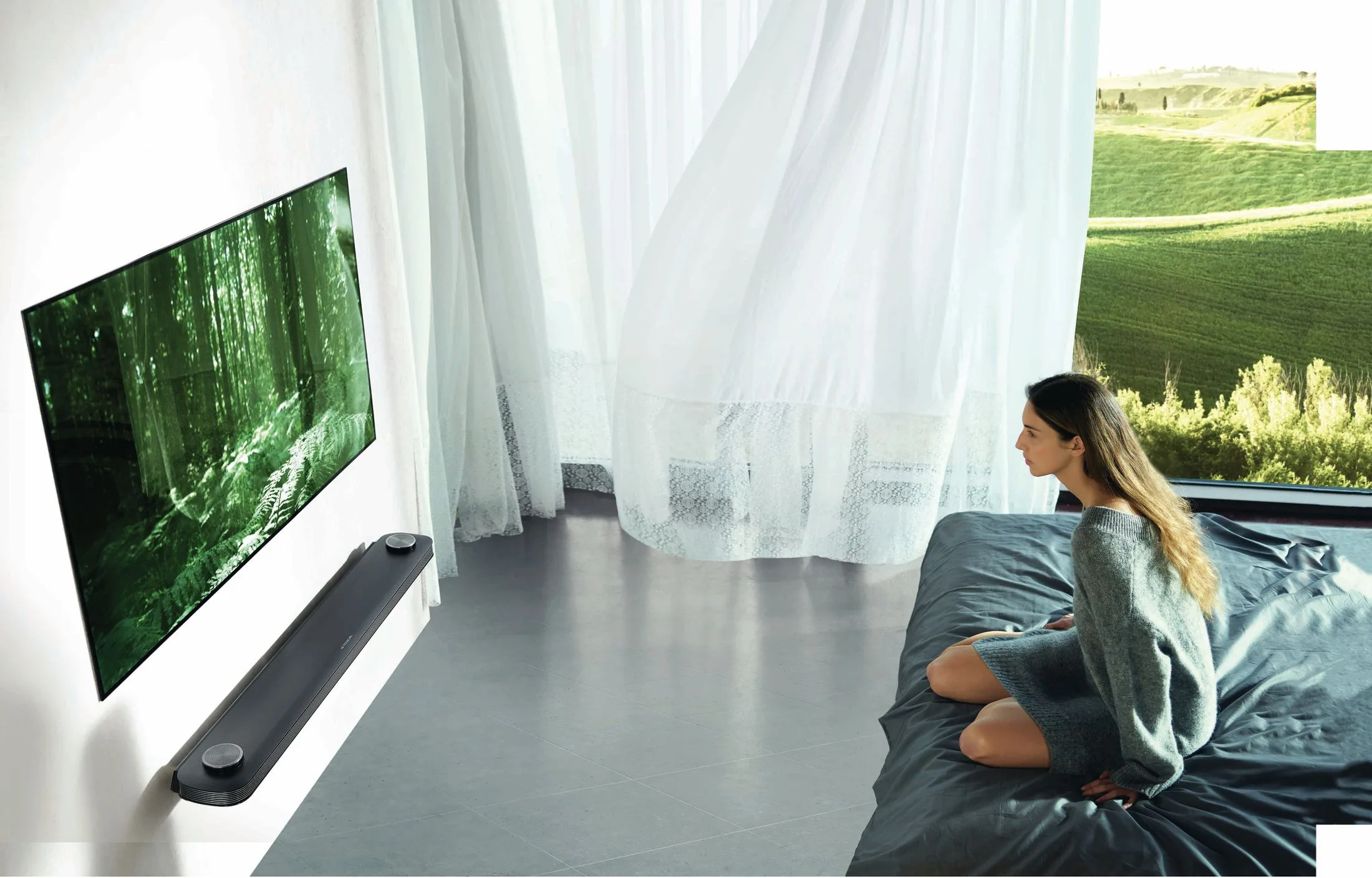 A woman watches a wall-mounted TV displaying a forest scene; sheer curtains and a scenic view are in the background.