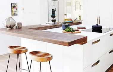 Modern kitchen with white cabinets, wooden bar, round stools, countertop decor, and wall art. Bright and stylish design.