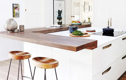 Modern kitchen with white cabinets, wooden bar, round stools, countertop decor, and wall art. Bright and stylish design.