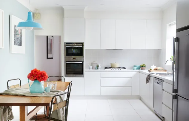 Bright, modern kitchen with white cabinets, dining table with red flowers, light blue pendant lamp, and stainless appliances.