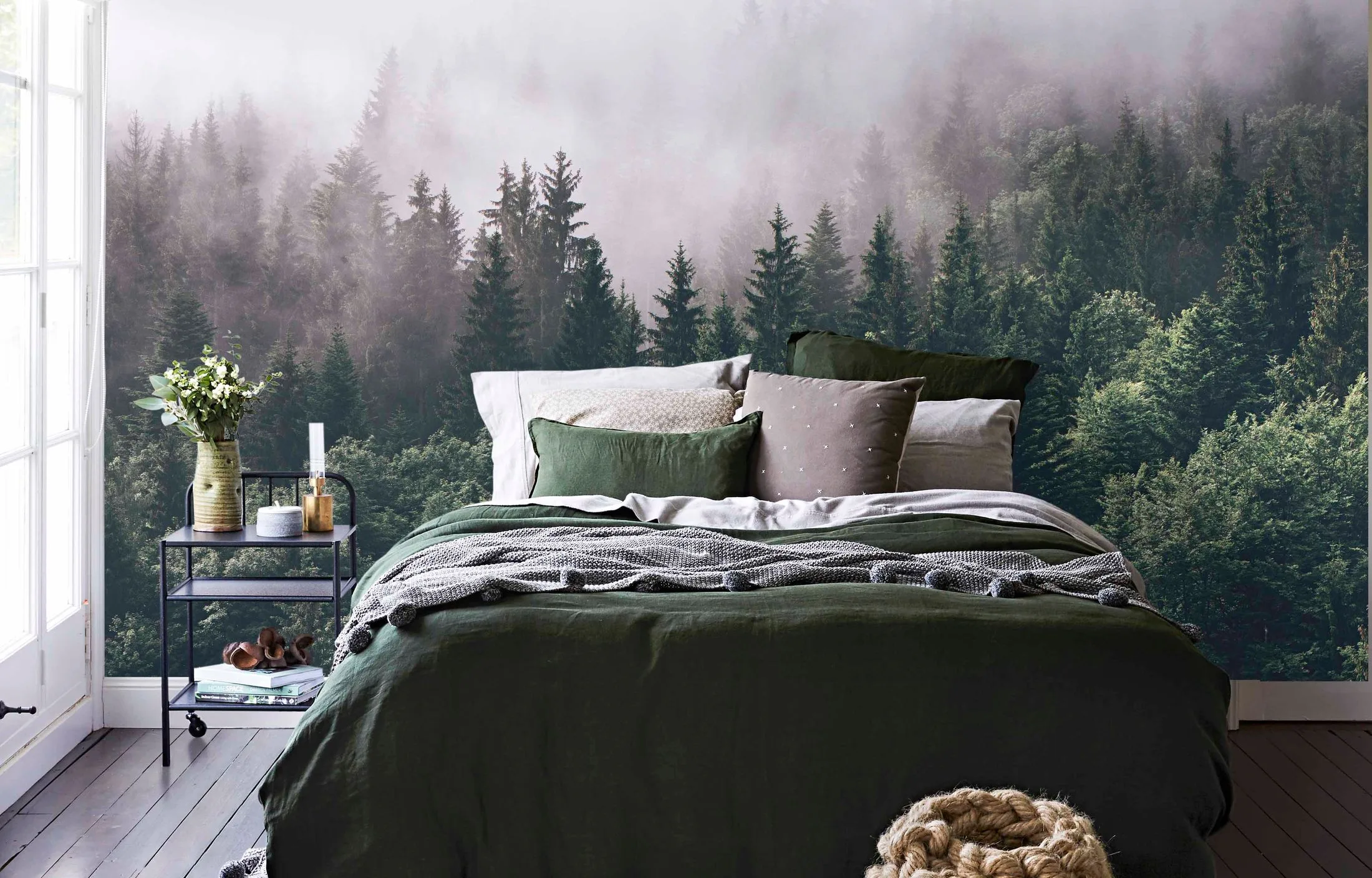 Cozy bedroom with a forest-themed wall, green bedding, and a nightstand with a vase and candle.
