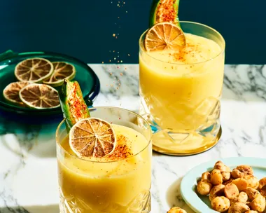 2 short tumbler style glasses with a mango liquid inside, garnished with a green jalapeno and dried lime. A ladies hand is hovering above squeezing lime into the cocktails. The cocktails are on a marble table top and navy blue background.