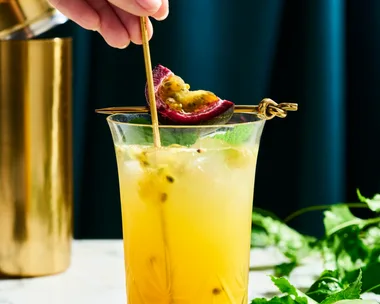a tall glass with yellow liquid in it with passionfruit seeds swirling in the liquid. This is on a marble table top with a dark navy background. You can find a cut slice of passionfruit next to the glass on the right side, as well as, passionfruit vine behind the glass.