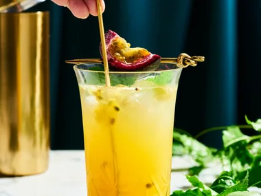 a tall glass with yellow liquid in it with passionfruit seeds swirling in the liquid. This is on a marble table top with a dark navy background. You can find a cut slice of passionfruit next to the glass on the right side, as well as, passionfruit vine behind the glass.