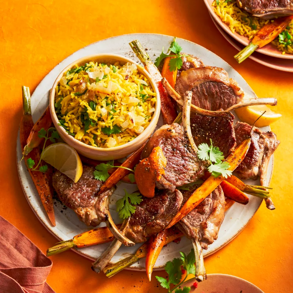lamb cutlets thrown on baby carrots with small bowl on plate with rice in it