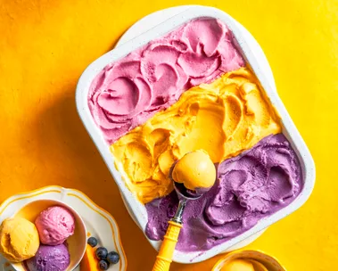 three flavours of gelato with purple, yellow and pink in one container with spoon scooping on top