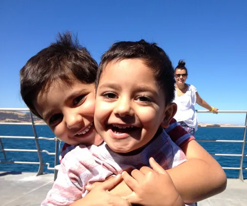 Two young boys smiling and hugging on a boat with an adult woman behind them, the sea and clear sky in the background.