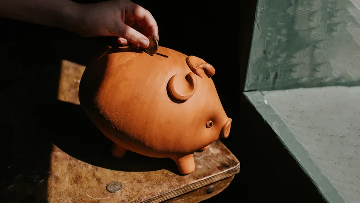 Hand inserting a coin into a clay piggy bank on a wooden table near a window.