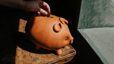 Hand inserting a coin into a clay piggy bank on a wooden table near a window.