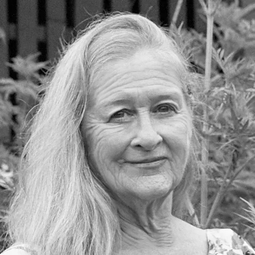 A woman with long hair smiles outdoors, surrounded by plants, in a black and white photograph.