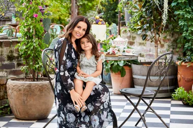 A woman and child smiling, sitting in a garden setting with flowers and a table with tea set and cake.