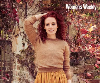 A woman with red curly hair in a brown sweater and yellow skirt poses outdoors in front of a rustic stone wall.