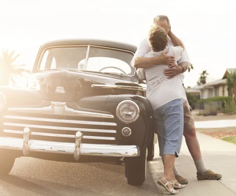 An elderly couple embraces next to a vintage car on a sunny street, with suburban homes and palm trees in the background.