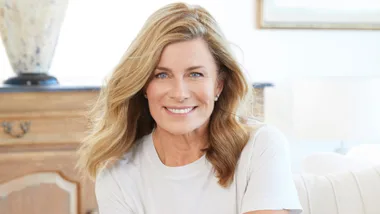 Smiling woman with wavy blonde hair in a white shirt, sitting indoors.