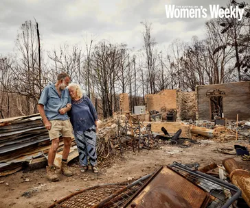 An elderly couple embraces, walking through the burnt remains of their home in a landscape devastated by fire.
