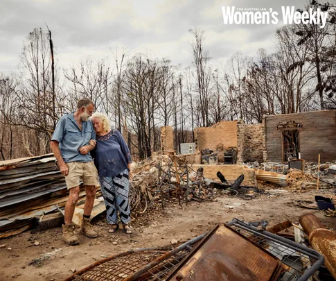 An elderly couple embraces, walking through the burnt remains of their home in a landscape devastated by fire.