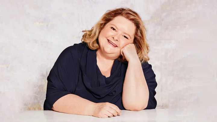 Smiling person with shoulder-length hair wearing a dark top, resting face on hand against a light background.