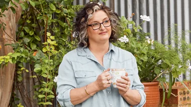 Woman in denim shirt holding a mug, standing outdoors in a lush garden setting.